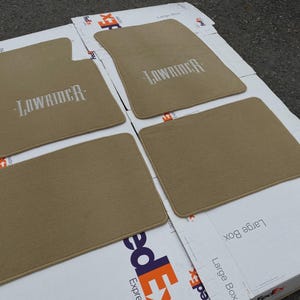 以下が含まれることがあります： 車両用のベージュのカーペットフロアマット4枚セット。2枚のマットには白い糸で「LOWRIDER」という文字が刺繍されています。マットは角が丸く、縁取りが施されており、自動車の車内フロアにフィットするように設計されています。
