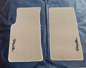 May include: A pair of light beige automotive floor mats with a contoured shape on one side. Each mat features the word Miata embroidered in black script lettering near the bottom corner and is finished with a light beige fabric edge binding.