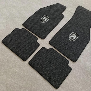 Puede incluir: Un juego de cuatro alfombrillas de coche de color gris oscuro con superficie texturizada. Dos de las alfombrillas presentan un emblema de escudo bordado en blanco que representa una puerta de castillo sobre agua, con formas personalizadas.