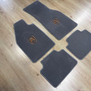 Puede incluir: Un juego de cuatro alfombrillas de coche de color gris oscuro con una textura de moqueta suave y bordes rematados. Dos de las alfombrillas presentan un emblema centrado en forma de escudo en tonos marrón y negro, mientras las otras dos son lisas.