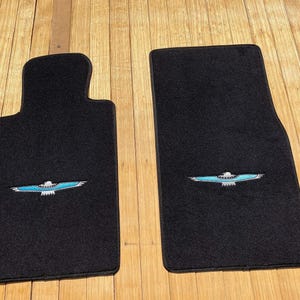 Può includere: Due tappetini per auto neri con un emblema ricamato al centro raffigurante un'aquila stilizzata con ali azzurre e corpo bianco. I tappetini hanno una superficie in moquette testurizzata e sono sagomati con bordi superiori arrotondati.