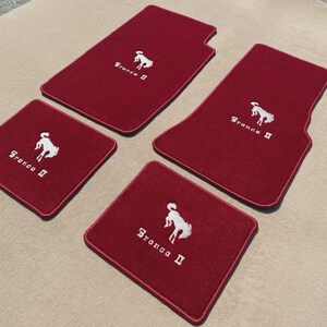Peut inclure: Un ensemble de quatre tapis de sol automobiles rouge foncé avec une bordure rouge assortie. Chaque tapis présente un motif brodé blanc représentant un cheval cabré et le texte Bronco II. Le lot comprend deux tapis avant et deux arrière.