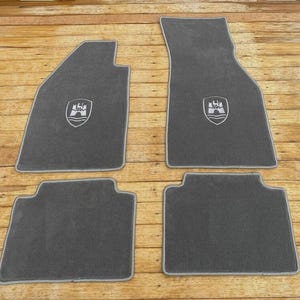 May include: A set of four dark gray automotive floor mats with light gray piping. Two front mats feature an embroidered crest depicting a castle above wavy lines, while the two rear mats are rectangular with notched corners and no embroidery.