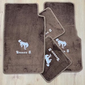 Peut inclure: Un ensemble de quatre tapis de sol automobiles marron avec un logo Bronco II brodé en blanc et un motif de cheval cabré. Les tapis présentent une texture en moquette épaisse avec des bords finis, conçus pour le plancher d'un véhicule.