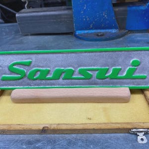 Könnte beinhalten: Ein rechteckiges Sansui-Logo-Emblem mit erhabener grüner Schrift auf strukturiertem silbernem Hintergrund, umrahmt von einem hellgrünen Rand. Das Emblem ist auf einem hellen Holzblock positioniert, der in einem flachen Holztablett liegt.