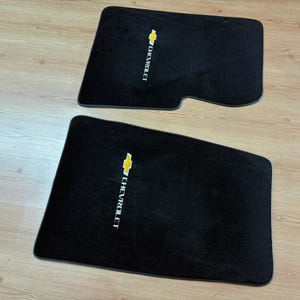 Puede incluir: Dos alfombrillas de coche negras con el logotipo de Chevrolet bordado en amarillo y blanco. Las alfombrillas tienen una textura de moqueta suave y están diseñadas con contornos específicos para el interior de un vehículo, mostrando la marca.