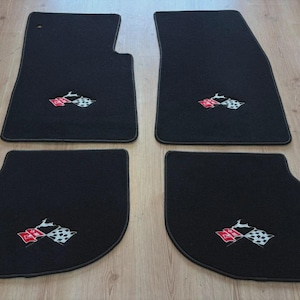Könnte beinhalten: Ein Set aus vier schwarzen Automatten mit einem gestickten Emblem aus zwei gekreuzten Flaggen, einer roten und einer karierten, mit einer kleinen weißen Tiersilhouette darüber. Die Matten haben abgerundete Ecken und einen dunklen Rand.