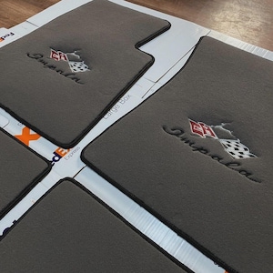 Op de afbeelding: Een set donkergrijze automatten met zwarte randen. Elke mat is voorzien van een geborduurd logo met het woord Impala in cursief schrift boven een afbeelding van twee gekruiste geblokte en rode vlaggen met een wit embleem in het midden.