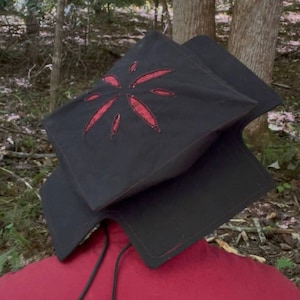 Könnte beinhalten: Eine schwarze Kopfbedeckung mit einem strukturierten, quadratischen Oberteil, das ein rotes, sternförmiges Ausschnittdesign mit genähten Kanten aufweist. Das Accessoire hat schwarze Kordeln und liegt auf einem einfarbigen roten Kleidungsstück.