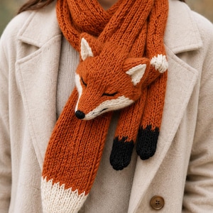 May include: A chunky knit scarf in a vibrant burnt orange color, featuring a three-dimensional fox head design. The fox has white accents on the ears and face, a black nose, and black paws at the ends of the scarf, with a white-tipped tail design.