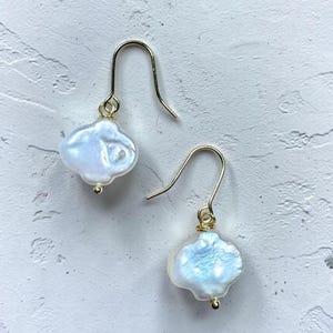 Puede incluir: Un par de pendientes colgantes con perlas de agua dulce blancas e iridiscentes de forma irregular, suspendidas de ganchos de metal dorado. Cada perla tiene una forma orgánica única con una superficie brillante y un pequeño abalorio dorado.
