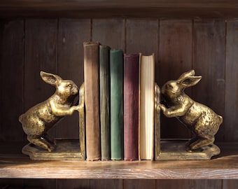 May include: A pair of gold-toned metal bookends, each shaped like a rabbit standing on its hind legs with front paws pressed against a flat vertical support. The two bookends hold a row of four hardcover books with brown, green, and maroon spines.