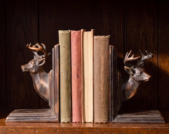 May include: A pair of dark brown bookends featuring sculpted deer heads with antlers, positioned on either side of four vintage books with muted green, red, and brown spines. The bookends have a smooth, metallic finish and L-shaped bases.