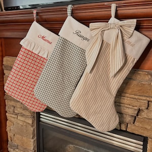 May include: Three Christmas stockings with cream-coloured cuffs. The first features a red and green plaid pattern with a ruffled trim and the name Mama. The second has a grey and white gingham pattern and the name Ranger. The third is beige striped.