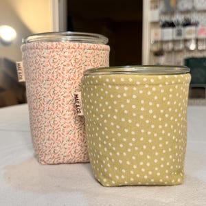 Peut inclure: Deux bocaux en verre avec des housses en tissu. Le plus grand bocal a un imprimé floral rose et le plus petit a un tissu vert clair à pois blancs. Chaque housse comporte une petite étiquette blanche avec le texte MAE & CO. cousu sur le côté.