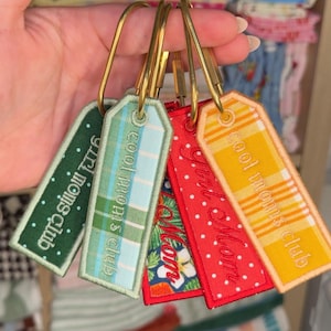 May include: Five rectangular fabric keychains with gold-tone metal rings. The tags feature embroidered text including "cool moms club" and "girl mom" in various patterns: green with white dots, light blue plaid, floral, red with white dots, and yellow plaid.