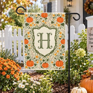 May include: A rectangular garden flag featuring a cream-coloured background with a central green shield containing the letter H. The design is surrounded by orange pumpkins, green vines, and small yellow flowers on a black metal stand.