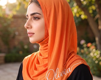 May include: A bright orange headscarf made of soft fabric, draped around the head and neck. The fabric features the name Olivia embroidered in a white cursive script font on the lower side, creating a clear contrast against the vibrant orange.
