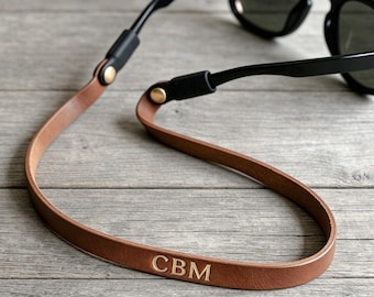 May include: A tan leather glasses strap attached to black sunglasses. The strap features gold-toned metal hardware at the connection points and the letters CBM embossed in gold on the leather surface, creating a functional accessory.