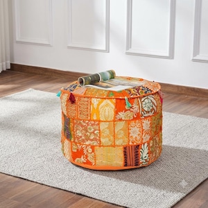 May include: A round, orange-toned floor pouf featuring a patchwork design of various embroidered fabric panels. The pouf is adorned with small, colorful tassels along the top edge and has a magazine resting flat on its surface.