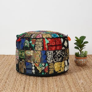 Puede incluir: Un puf de suelo redondo con un diseño de patchwork de varios tejidos bordados en negro, rojo, verde, azul y dorado. La superficie está decorada con intrincados patrones florales, hilos metálicos, pequeños espejos y borlas coloridas.