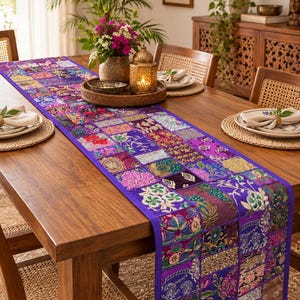 Pode incluir: Um caminho de mesa retangular longo com um design de retalhos de vários quadrados de tecido em tons de roxo, rosa, dourado e verde. O caminho é decorado com bordados intrincados, detalhes em fio metálico e padrões florais.
