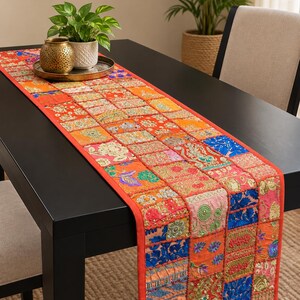 Può includere: Un runner da tavola rettangolare caratterizzato da un design patchwork di vari quadrati di tessuto in tonalità vivaci di arancione, rosso, blu e oro. Il runner è decorato con ricami complessi, fili metallici e piccole paillettes sulla superficie.