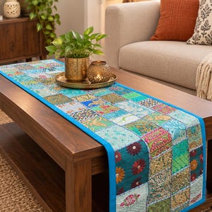 Peut inclure: Un chemin de table rectangulaire composé d'un assemblage de carrés de tissu dans des tons de bleu sarcelle, turquoise, vert et rose. Le chemin est orné de broderies complexes, de paillettes et de motifs floraux, avec une bordure bleue unie.