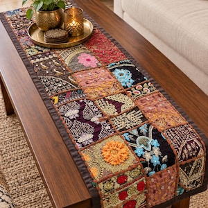 Peut inclure: Un chemin de table rectangulaire présentant un motif patchwork composé de divers carrés de tissu dans des tons de rouge, orange, violet, noir et bleu. Le chemin est orné de broderies, de paillettes et de perles, avec une bordure marron foncé.