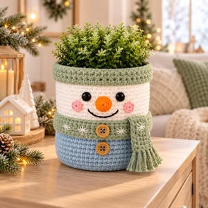 May include: A crochet snowman-shaped basket featuring a white face with black eyes, a bright orange nose, and pink cheeks. The basket has a sage green rim and a matching scarf with white snowflake embroidery and two wooden buttons.