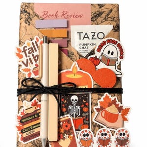 Puede incluir: Un set de regalo de libro envuelto que incluye una bolsita de té Tazo Pumpkin Chai, un bolígrafo, un resaltador y varias pegatinas de temática otoñal como fantasmas leyendo, calabazas y tazas de café. El paquete está atado con cordel negro.