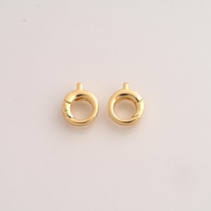 May include: Two gold-toned circular spring ring clasps, each featuring a small protruding tab at the top. These jewelry findings have a polished, reflective metallic surface and a circular opening in the center for connecting jewelry components.