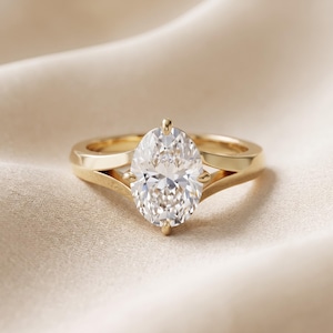May include: A gold ring featuring a large, clear, oval-cut center stone held by four prongs. The band is crafted from polished yellow gold and splits into two distinct sections as it reaches the central setting, creating a sleek, open design.