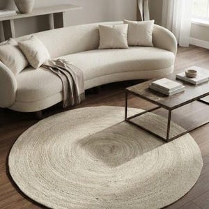 May include: A large, round, woven area rug in a neutral beige tone, featuring a textured, concentric circular pattern. The rug is placed on a dark wood floor in front of a curved, cream-colored sofa with matching throw pillows and a throw blanket.