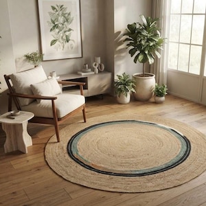 May include: A round, woven jute rug featuring a natural tan base with concentric circular bands in shades of blue, teal, and dark gray. The rug is positioned on a light-colored wooden floor in a room with a chair, plants, and furniture.