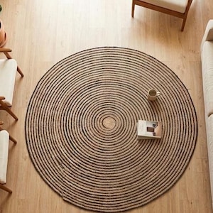 May include: A large round area rug featuring a concentric circular pattern in alternating dark brown and natural tan fibers, positioned in the center of a room with a beige sofa, two wooden armchairs, and a small potted plant.