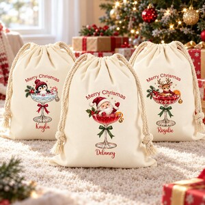May include: Three off-white canvas drawstring gift bags featuring festive illustrations of a penguin, Santa Claus, and a reindeer inside cocktail glasses. Each bag displays the text Merry Chrismas and a personalised name: Kayla, Delaney, and Rosalie.