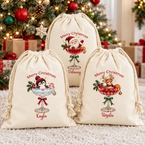 May include: Three off-white canvas drawstring gift bags featuring Christmas graphics. Each bag displays a cocktail glass filled with a character: Santa, a penguin, or a reindeer. Text reads Merry Christmas and names Delaney, Kayla, Rosalie.