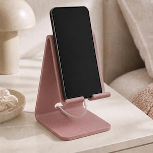 Könnte beinhalten: Ein altrosa-farbener, L-förmiger Handyhalter mit matter Oberfläche und einer kleinen Aussparung für ein Ladekabel. Ein schwarzes Smartphone steht vertikal auf dem Halter, wobei ein weißes Ladekabel an der Unterseite des Geräts eingesteckt ist.