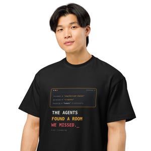 以下が含まれることがあります： 黒の半袖Tシャツ。コンピューターのターミナルウィンドウのグラフィックが描かれており、コードテキストと「THE AGENTS FOUND A ROOM WE MISSED.」というフレーズがプリントされています。文字は黒い生地の上に白、黄色、赤で表示されています。