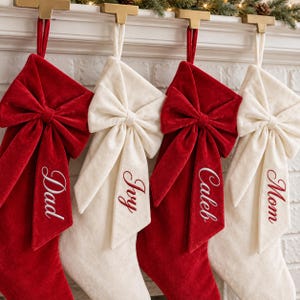May include: Four velvet Christmas stockings hanging in an alternating red and cream color pattern. Each stocking features a large, matching velvet bow at the top and is embroidered with a name: Dad, Ivy, Caleb, and Mom in script font.