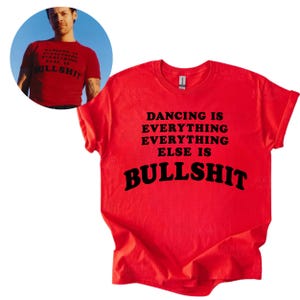 May include: A bright red short-sleeved t-shirt featuring black text that reads DANCING IS EVERYTHING EVERYTHING ELSE IS BULLSHIT. The shirt has a crew neckline and is displayed flat, with a circular inset showing the shirt worn by a person.