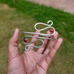 May include: A silver-toned metal cuff bracelet featuring a swirling, open-ended design. The bracelet is accented with two small, round, polished red stones, each set at the terminal end of the curved metal wire structure.