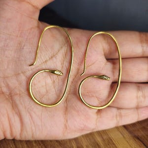 Könnte beinhalten: Ein Paar goldfarbene Metallohrringe in Form von länglichen Schlaufen mit einem Schlangenkopf-Detail am Ende jedes Drahtes. Die Ohrringe haben eine glatte, polierte Oberfläche und ein geschwungenes Design, das der Kontur des Ohrs folgt.