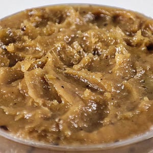 May include: A thick, textured paste with a golden-brown hue, featuring visible small dark specks throughout the mixture. The surface displays uneven, swirled peaks and valleys, creating a dense, semi-solid consistency with a slight sheen.
