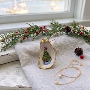 May include: A decorative oyster shell ornament featuring a hand-painted green Christmas tree with a red star topper, finished with a gold-painted rim and a thin twine loop for hanging, accompanied by a delicate gold chain necklace with a pearl.