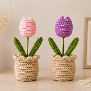 Op de afbeelding: Twee gehaakte tulpen in beige potten. De ene tulp heeft een lichtroze bloem, de andere een paarse bloem. Beide hebben groene stelen en twee groene bladeren die uit de bovenkant van de beige garen potten met geschulpte rand komen.