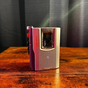 May include: A silver and purple Sony cassette recorder featuring a speaker grille, a clear window for the cassette, and the text "SONY" and "CLEAR VOICE" printed on the front. The device includes a red button and several black control buttons.