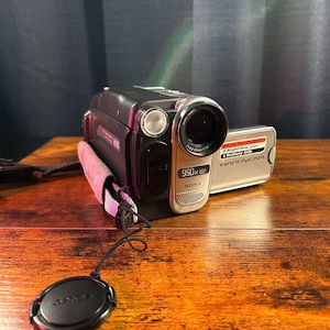 Op de afbeelding: Een zwart-zilveren Sony Handycam camcorder met een 20x optische zoomlens, 990x digitale zoom en NightShot Plus-functie. Het apparaat is voorzien van een roze en zwarte handriem, een losse zwarte lensdop en een display aan de zijkant.