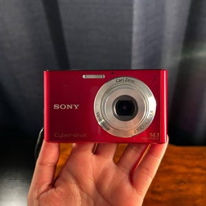 Puede incluir: Una cámara digital Sony Cyber-shot de color rojo con un objetivo Carl Zeiss Vario-Tessar. La parte frontal muestra el logotipo de Sony, la marca Cyber-shot y el texto 14.1 Mega Pixels impreso en la parte inferior derecha del cuerpo.